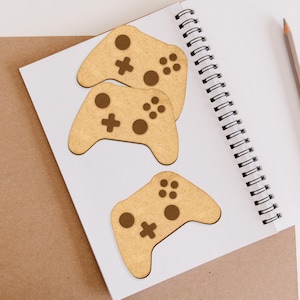 May include: Three wooden game controller cutouts in brown with brown details. The cutouts are on a white notebook with a spiral binding.