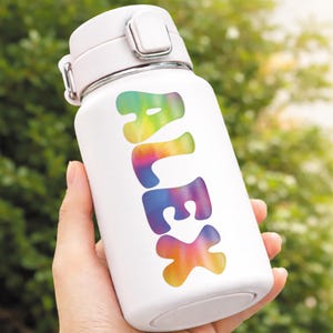 May include: White insulated water bottle with a flip-top lid. The word "ALEX" is printed vertically in a tie-dye rainbow design. The bottle is held in a person's hand against a blurred green background.