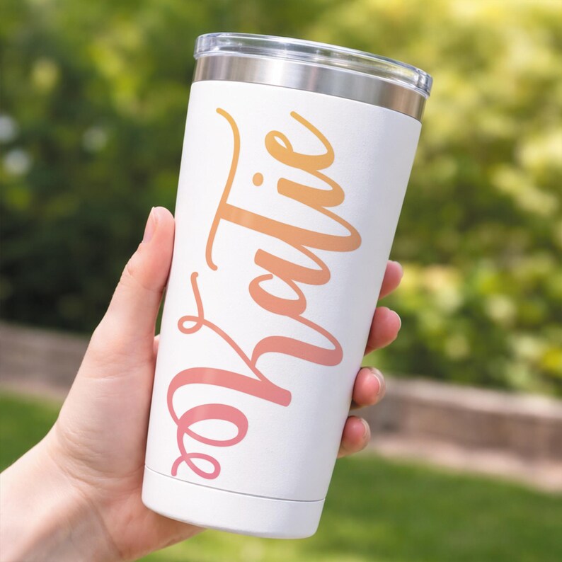 May include: White insulated tumbler with a clear lid, held by a hand. The tumbler features the name "Katie" in a gradient of orange and pink script. The background is blurred green foliage.