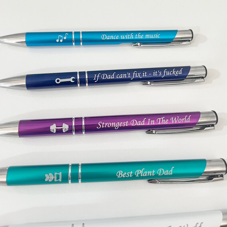 May include: Four different colored pens with silver accents and engraved text. The pens are blue, navy blue, purple, and teal. The text on the pens reads "Dance with the music", "If Dad can't fix it - it's fucked", "Strongest Dad In The World", and "Best Plant Dad".