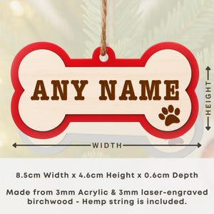 Peut inclure: Ornement en forme d'os de chien avec une bordure rouge et un centre en bois clair. Le texte "ANY NAME" est affich&eacute; en lettres marron. Une empreinte de patte est sur le c&ocirc;t&eacute; droit. L'ornement mesure 8,5 cm de large et 4,6 cm de haut.