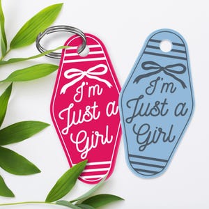 K&ouml;nnte beinhalten: Zwei Schl&uuml;sselanh&auml;nger mit dem Text "I'm Just a Girl". Einer ist rosa mit wei&szlig;en Details, der andere hellblau mit grauen Details. Beide haben eine Schleife und sind an einem silbernen Schl&uuml;sselring befestigt. Retro-Motel-Stil.