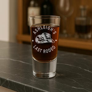 K&ouml;nnte beinhalten: Ein klares Schnapsglas, gef&uuml;llt mit einer braunen Fl&uuml;ssigkeit, mit dem wei&szlig;en Text "Ashleigh's Last Rodeo" und einer Cowboyhut-Grafik. Das Glas steht auf einer dunklen Arbeitsplatte. Der Hintergrund ist verschwommen.