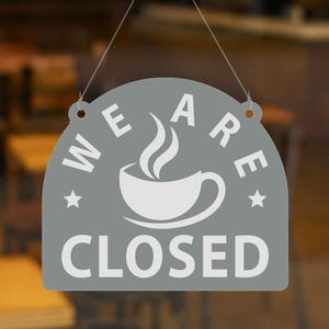 May include: A gray, semi-circular sign with the words "WE ARE CLOSED" in white lettering. A white coffee cup with steam is in the center. The sign is hanging by a thin string.