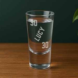 May include: A clear shot glass filled with liquid, with the name "LUCY" in white, and the numbers "30" above and below. The glass is set against a dark green background on a wooden surface.