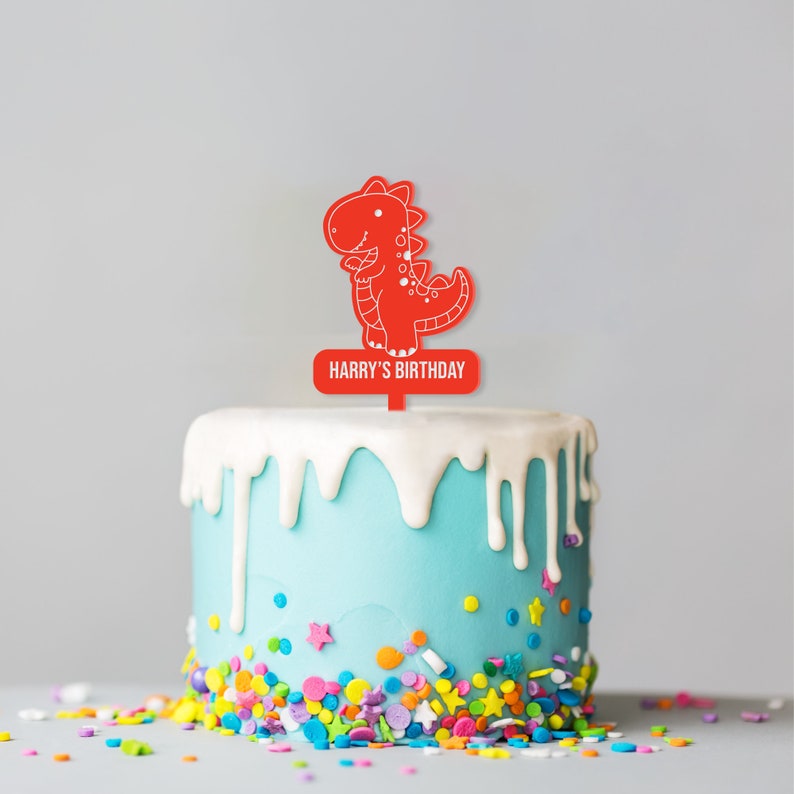 May include: A red dinosaur cake topper with the text "Harry's Birthday" on a blue cake with white frosting and colorful sprinkles.
