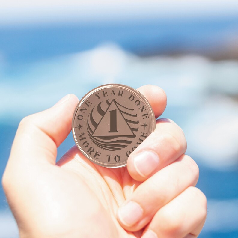 Op de afbeelding: Een bronzen munt met de woorden "One Year Done" en "More To Come" rond de rand. Het midden van de munt heeft een driehoek met het nummer "1" erin en een golvend ontwerp.