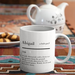 May include: White ceramic mug with a black definition of the name Abigail, pronounced "a-buh-gayl". The definition reads "Overly distracted, with a keen eye for pennies from the ground. With a overly obsession for cats and board games. Widely loved and hated by many."