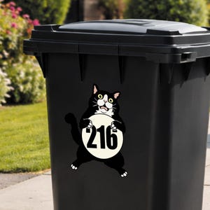 May include: Black trash bin with a cartoon cat sticker. The cat is black and white, holding a white circle with the number 216 in black. The bin is outdoors, with green grass and bushes in the background.