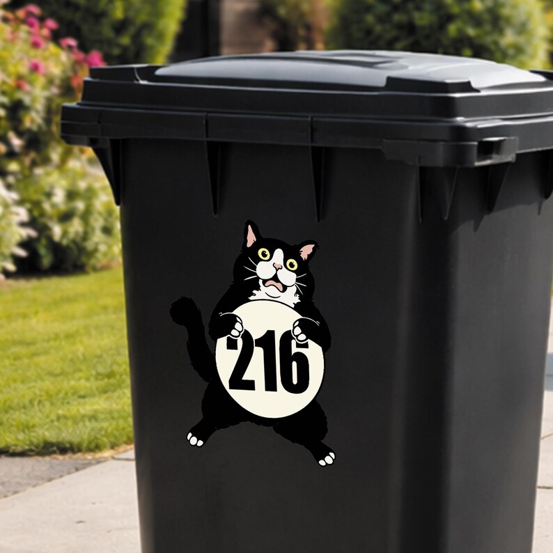 May include: Black trash bin with a cartoon cat sticker. The cat is black and white, holding a white circle with the number 216 in black. The bin is outdoors, with green grass and bushes in the background.
