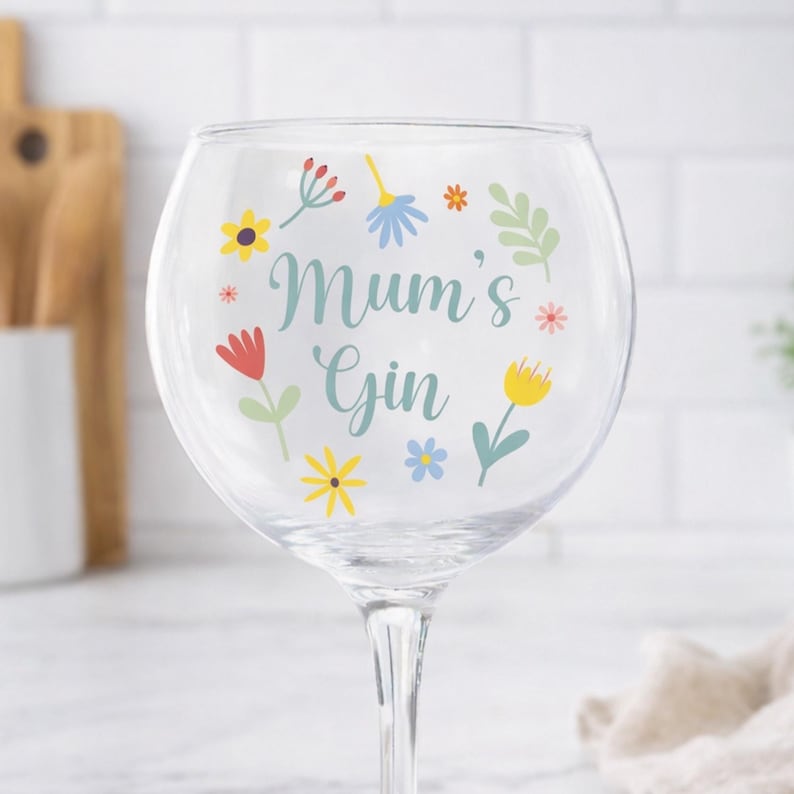 Może przedstawiać: Przezroczysty kieliszek do ginu z kwiatowym wzorem i napisem "Mum's Gin". Kwiaty są w odcieniach ż&oacute;łci, czerwieni, niebieskiego i zieleni. Kieliszek ma długą n&oacute;żkę.