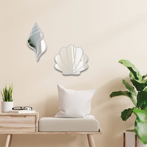May include: Two decorative wall mirrors shaped like seashells. The larger mirror is a scallop shell and the smaller mirror is a conch shell. Both mirrors are silver and have a reflective surface.
