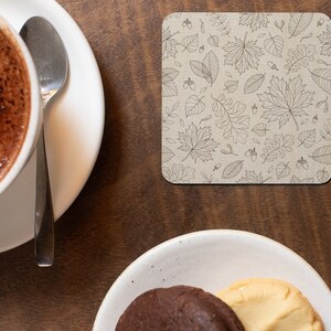 May include: A close-up of a brown wooden table with a white ceramic cup of coffee and a silver spoon on a white saucer. A square coaster with a black and white leaf pattern is on the table. A white ceramic plate with two cookies is on the table.
