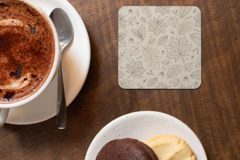 May include: A close-up of a brown wooden table with a white ceramic cup of coffee and a silver spoon on a white saucer. A square coaster with a black and white leaf pattern is on the table. A white ceramic plate with two cookies is on the table.