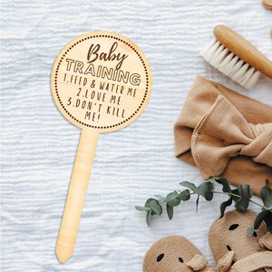 May include: A light-colored wooden sign with the words "Baby TRAINING" and instructions: "1. FEED & WATER ME 2. LOVE ME 3. DON'T KILL ME!" The sign is round with a dotted border and a handle. Other baby items are in the background.