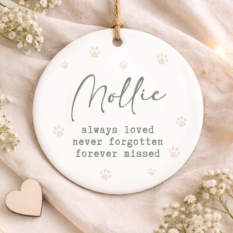 May include: A white ceramic pet memorial ornament with the name "Mollie" in script, surrounded by paw prints. The text "always loved, never forgotten, forever missed" is below the name. A jute string is attached for hanging.