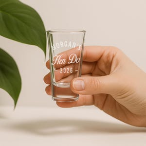 May include: A clear glass shot glass with the text "Morgan's Hen Do - 2026" in a white script font. The shot glass is held by a person's hand, with a green leaf in the background.
