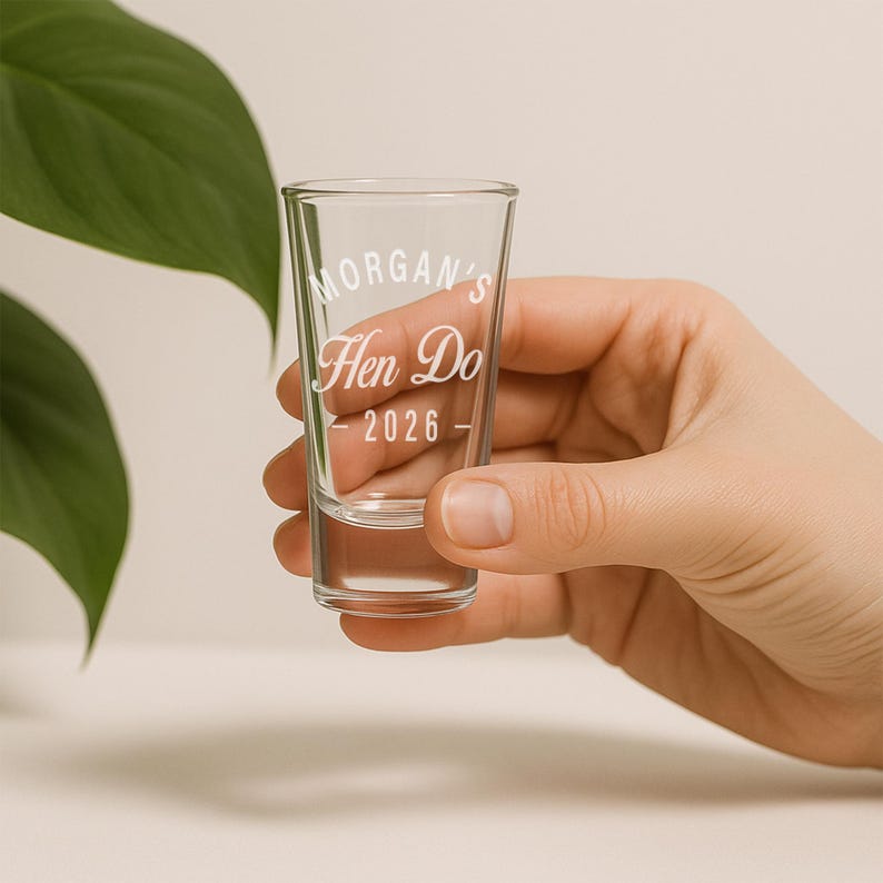 May include: A clear glass shot glass with the text "Morgan's Hen Do - 2026" in a white script font. The shot glass is held by a person's hand, with a green leaf in the background.