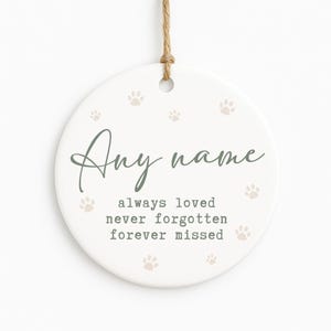 May include: A white, round ceramic ornament with a jute twine hanger. The ornament features the text "Any name" in a script font, along with the phrases "always loved, never forgotten, forever missed." Small paw prints are scattered across the surface.