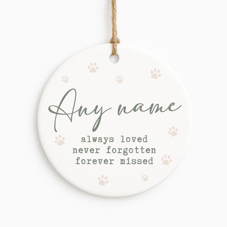 May include: A white, round ceramic ornament with a jute twine hanger. The ornament features the text "Any name" in a script font, along with the phrases "always loved, never forgotten, forever missed." Small paw prints are scattered across the surface.