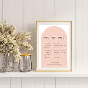 May include: A framed print with a gold border displays "OPENING TIMES" in a minimalist design. The print lists business hours for a coffee shop, with a peach-colored arch at the top. A vase of dried flowers and a glass jar are on the shelf.