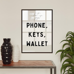 May include: A black framed print with white text that reads "Don't forget PHONE, KEYS, WALLET" on a white background. The print is hanging on a white wall above a wooden shelf with two black and white vases.