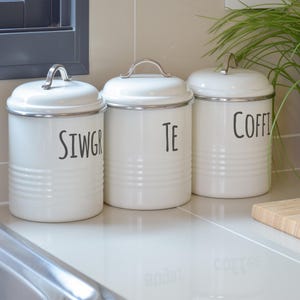 May include: Three white ceramic canisters with black lettering. The canisters are labeled "Sugar", "Tea", and "Coffee".