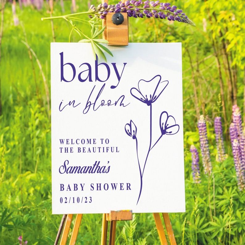 May include: White sign with purple text and a purple flower illustration. The text reads "baby in bloom" and "Welcome to the beautiful Samantha's Baby Shower 02/10/23".