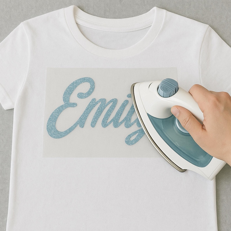May include: A white t-shirt with the name "Emily" in light blue glitter lettering being ironed on. A hand holds a white and blue iron over the transfer paper. The shirt is on a gray surface.