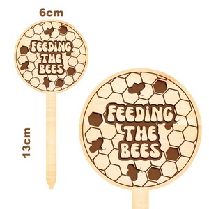 K&ouml;nnte beinhalten: Holz-Gartenpfahl mit einem runden, hellbraunen Schild, auf dem in brauner Schrift "F&uuml;ttern der Bienen" steht. Das Schild hat ein Wabenmuster mit kleinen Bienensilhouetten. Der Pfahl ist 13 cm hoch und 6 cm breit.