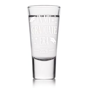 May include: Clear shot glass with the text "IN MY GRADUATE ERA 2026" in white. The glass has a cylindrical shape and is filled with a clear liquid. Graduation cap and star graphics are also included.