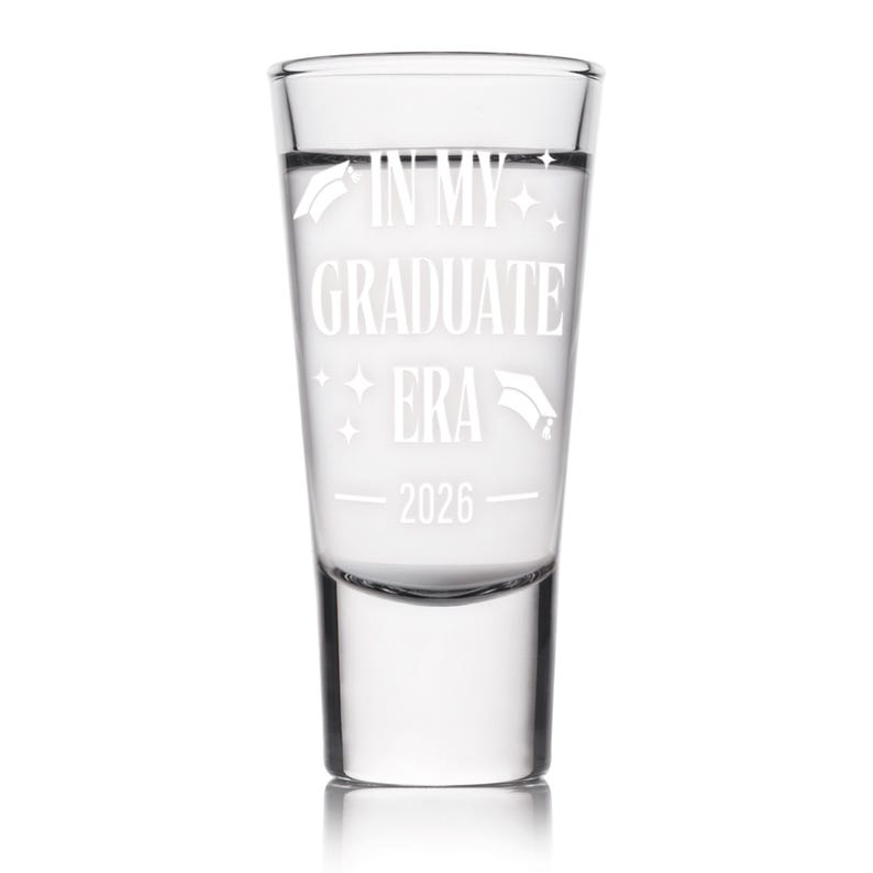 May include: Clear shot glass with the text "IN MY GRADUATE ERA 2026" in white. The glass has a cylindrical shape and is filled with a clear liquid. Graduation cap and star graphics are also included.