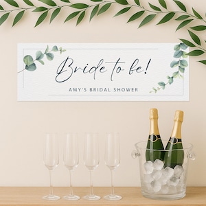 May include: A rectangular sign with the text "Bride to be!" and "Amy's Bridal Shower" in elegant script. The sign is decorated with watercolor eucalyptus leaves. Below, four champagne flutes and two champagne bottles in an ice bucket.