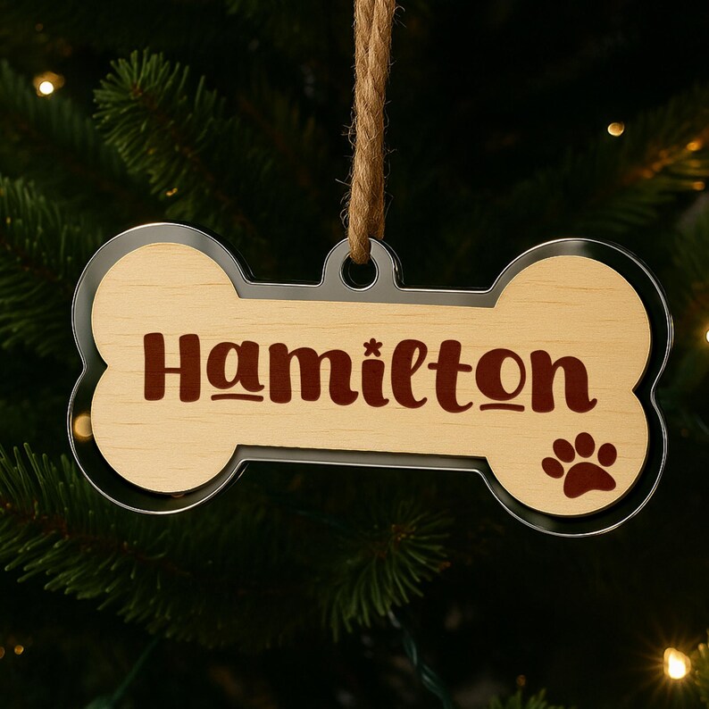 May include: A bone-shaped Christmas ornament with the name "Hamilton" in a dark brown script. The ornament has a light wood grain center, a paw print, and a silver-colored border. It hangs from a natural twine string.