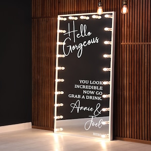 May include: A large, rectangular mirror with a white frame and a black interior. The mirror is surrounded by small, round lights. White script reads "Hello Gorgeous" and a message below. The mirror is leaning against a wooden wall.