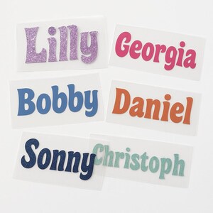 May include: Six iron-on name labels with different names. The names are Lilly, Georgia, Bobby, Daniel, Sonny, and Christoph. The labels are on a white background.