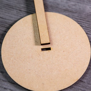May include: A round, light brown wooden base with a small slot cut into the top. A thin, rectangular piece of wood is placed on top of the base.