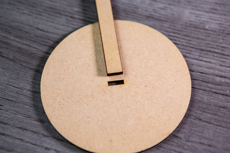 May include: A round, light brown wooden base with a small slot cut into the top. A thin, rectangular piece of wood is placed on top of the base.