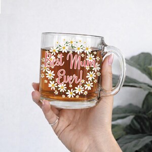 Puede incluir: Taza de vidrio transparente llena de t&eacute;, con el texto rosa "Best Mum Ever!" y margaritas blancas. La taza tiene un asa y est&aacute; sostenida por una mano.