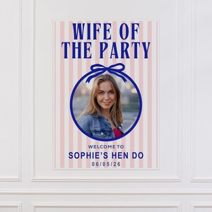 Puede incluir: Un p&oacute;ster vertical con el texto "WIFE OF THE PARTY" en letras may&uacute;sculas azules. Una foto de una mujer est&aacute; en un c&iacute;rculo azul con un lazo. Debajo de la foto est&aacute; "WELCOME TO SOPHIE'S HEN DO 06/05/26" sobre un fondo a rayas rosas y blancas.