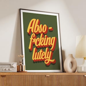 Puede incluir: Impresi&oacute;n art&iacute;stica enmarcada con la frase "Abso-f*cking-lutely" en letras 3D amarillas y rojas sobre un fondo verde oscuro. La impresi&oacute;n est&aacute; en un marco negro y se muestra en un mueble de madera con objetos decorativos.
