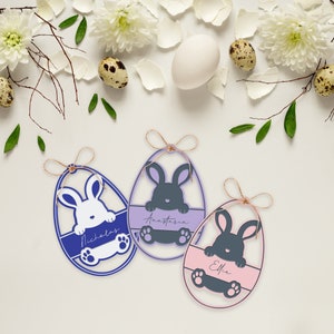 May include: Three Easter egg-shaped ornaments with bunny designs and personalized names. The eggs are in blue, purple, and pink, with a bunny peeking over a horizontal bar. Each egg is adorned with a twine bow.