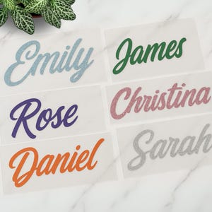 May include: Five personalized name labels with glitter lettering. The names are Emily, James, Christina, Sarah, and Daniel. The labels are on a white background.