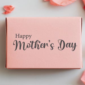 Puede incluir: Una caja de regalo rosa con el texto "Happy Mother's Day" impreso en la parte delantera.