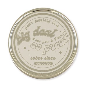 May include: A round, silver-colored coin with a beige background. The coin features the text "your sobriety is a big deal I see you & I am so proud sober since 00/00/00".