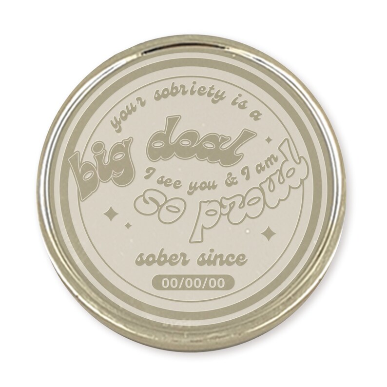 May include: A round, silver-colored coin with a beige background. The coin features the text "your sobriety is a big deal I see you & I am so proud sober since 00/00/00".
