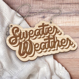 May include: A wooden sign with the words "Sweater Weather" in a brown, stylized font. The sign is light brown with a darker brown outline and is hanging by a string. The sign is resting on a white knitted blanket, with a wooden background.