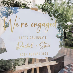 May include: A white acrylic sign with gold lettering announcing an engagement. The sign reads "We're engaged" with a gold brushstroke design. Below the text, it says "Let's celebrate Rachel + Steve 30th August 2023". The sign is displayed on a wooden easel.