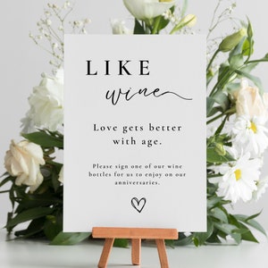 May include: A white sign with black text that reads "LIKE wine Love gets better with age. Please sign one of our wine bottles for us to enjoy on our anniversaries." A small heart is drawn at the bottom of the sign.