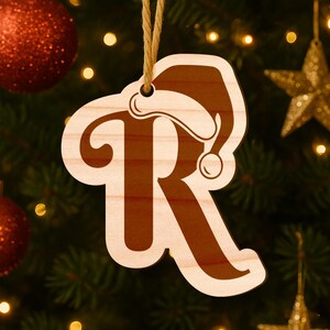 May include: Wooden Christmas ornament shaped like the letter "R" with a Santa hat design. The ornament is light brown with a darker brown interior and a beige border. It hangs from a natural-colored string.